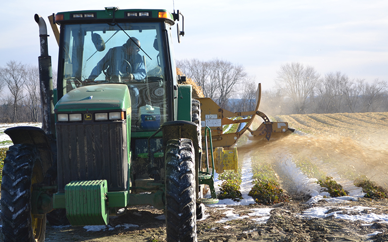 A man driving large green tractor with a plow driving through  a field of turned soil and some snow covering