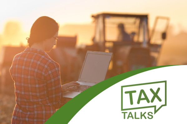 Young woman working on a lap top computer standing in an agriculture field in front of a tractor. Tax Talk logo is in the lower right hand corner.