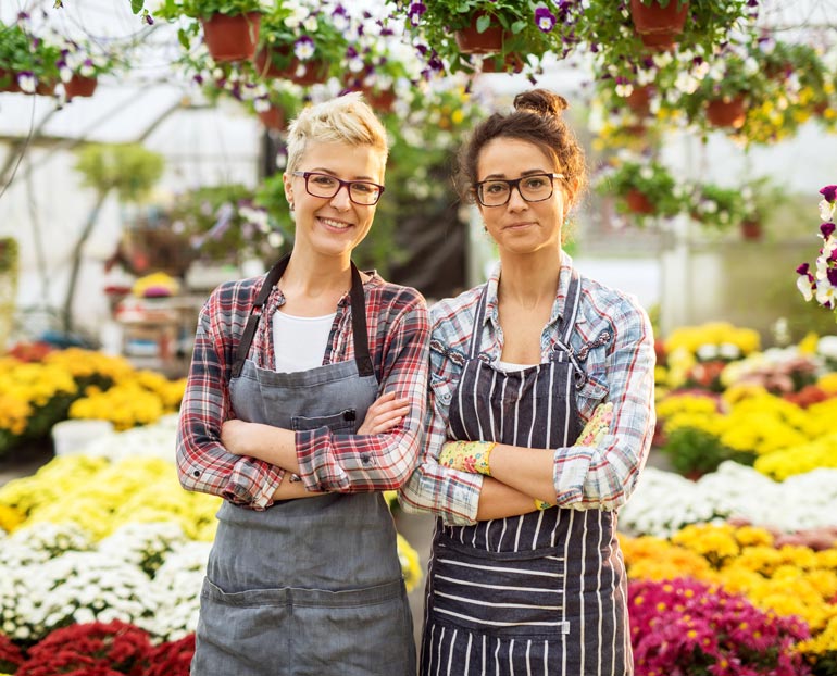 Two females stand cross-armed in greenhouse full of flowers 