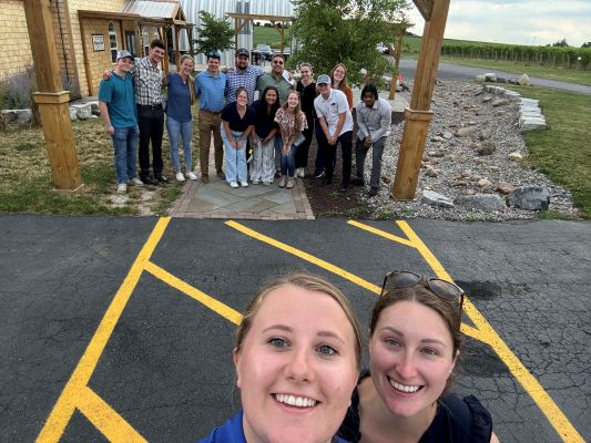 Two people talking a selfie with a group of people in the backl