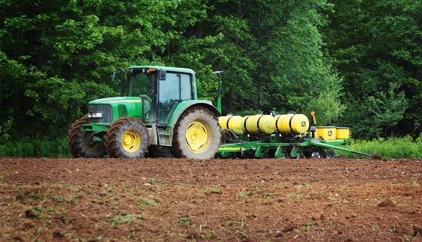 Green tractor pulling a yellow row planter across a freshly tilled field, with a dense tree line in the background under daylight conditions.