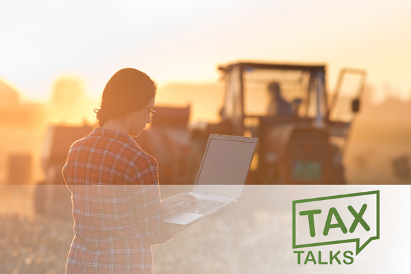 Woman in front of tractor in agriculture field holding and looking at a laptop computer, with tax talk logo in corner