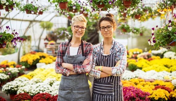 Two females stand cross-armed in greenhouse full of flowers  